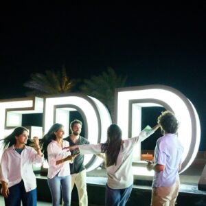 Wide shot of friends dancing and celebrating together near the illuminated Jeddah Corniche sign, enjoying fun travel