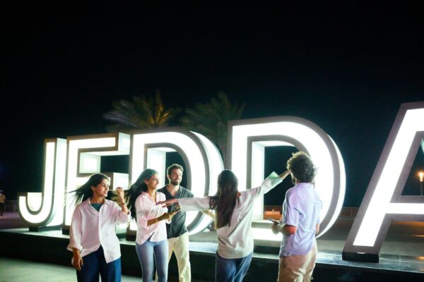 Wide shot of friends dancing and celebrating together near the illuminated Jeddah Corniche sign, enjoying fun travel